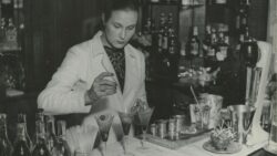 Valkotakkinen baarimestari sekoittaa drinkkejä mustavalkoisessa kuvassa.